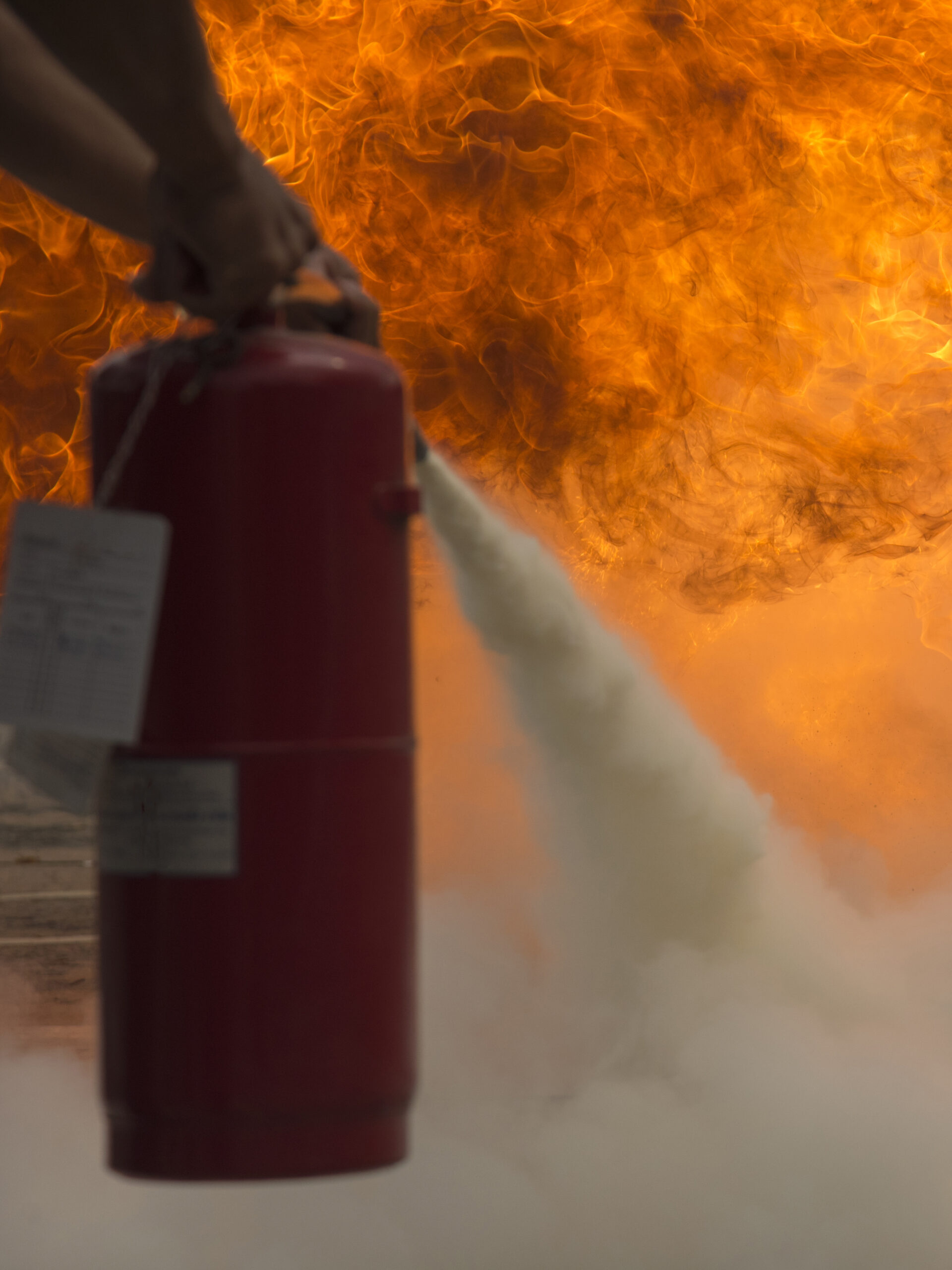 Gros plan sur une main tenant un extincteur rouge, projetant un jet de poudre ou de gaz vers un feu intense en arrière-plan avec de grandes flammes orange.