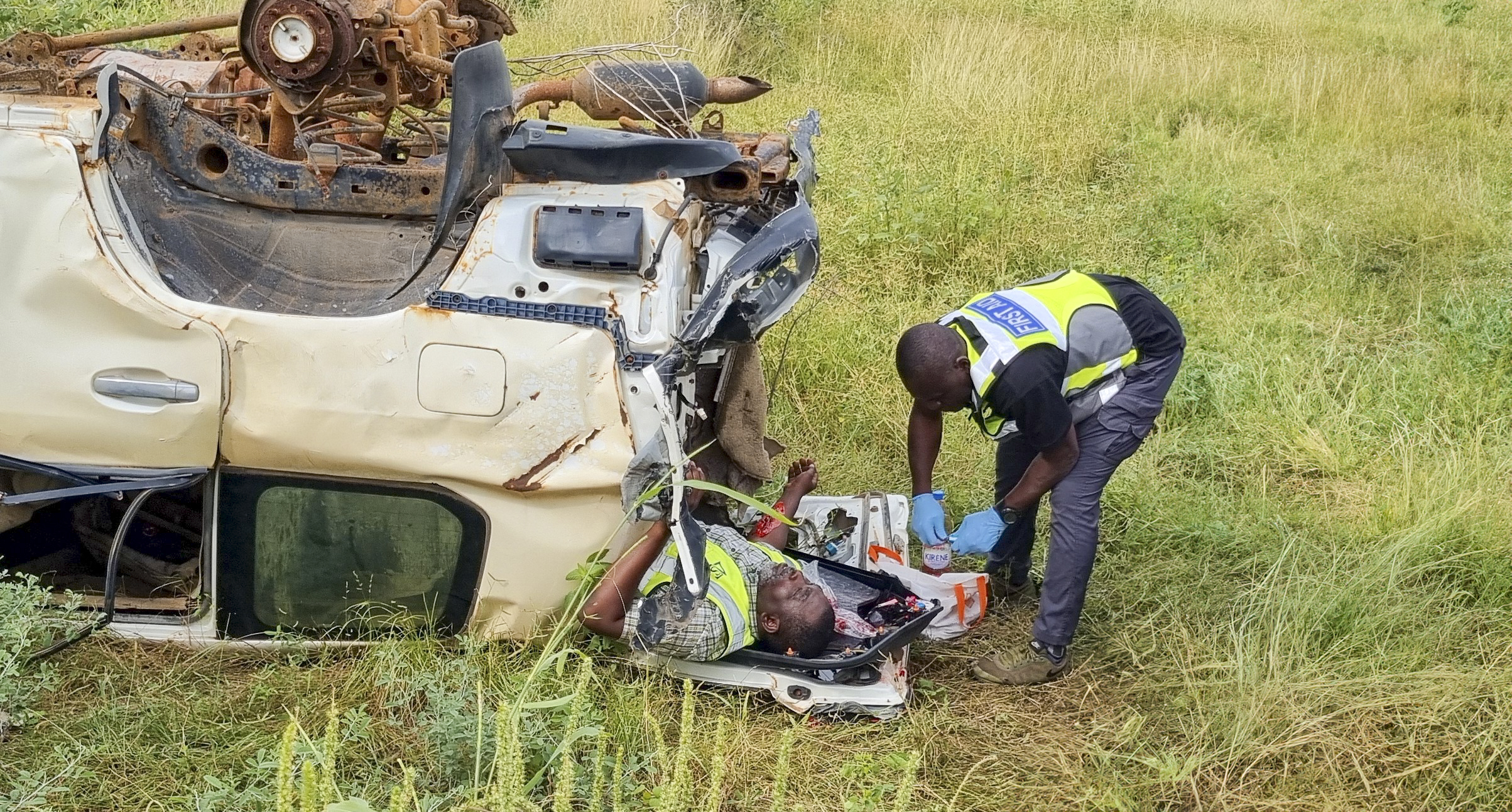 AACCES - Secouriste AACCES portant assistance à une victime après un accident de la route, à côté d'un véhicule retourné et endommagé, dans un environnement herbeux.